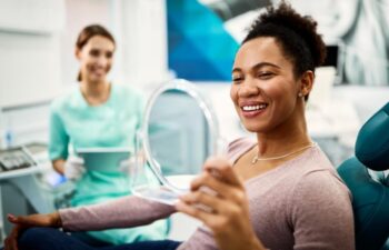 Satisfied African American woman using mirror while checking her teeth after appointment at dentist's office.