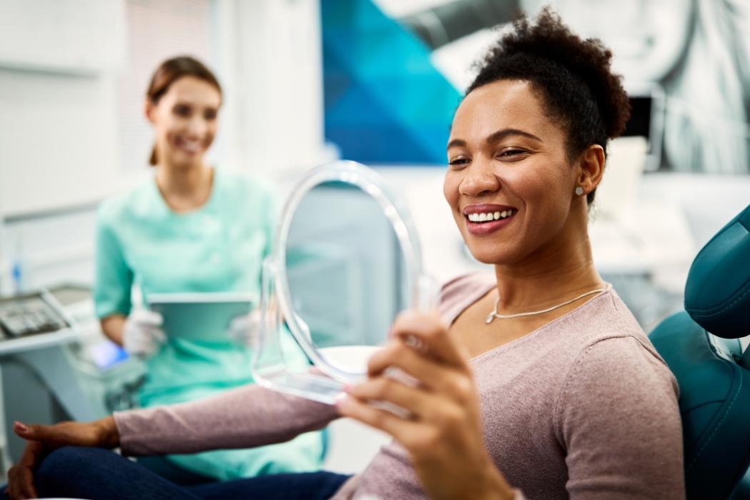 Satisfied African American woman using mirror while checking her teeth after appointment at dentist's office.