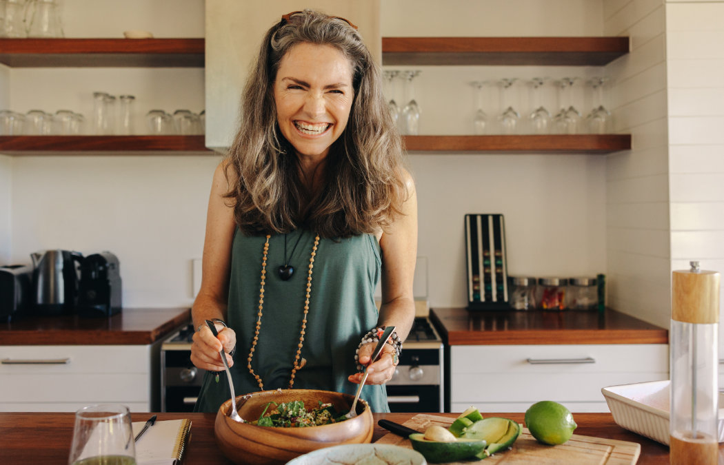 A woman is smiling while preparing food in a kitchen, holding utensils over a wooden bowl. Ingredients like avocado and lime are on the counter.