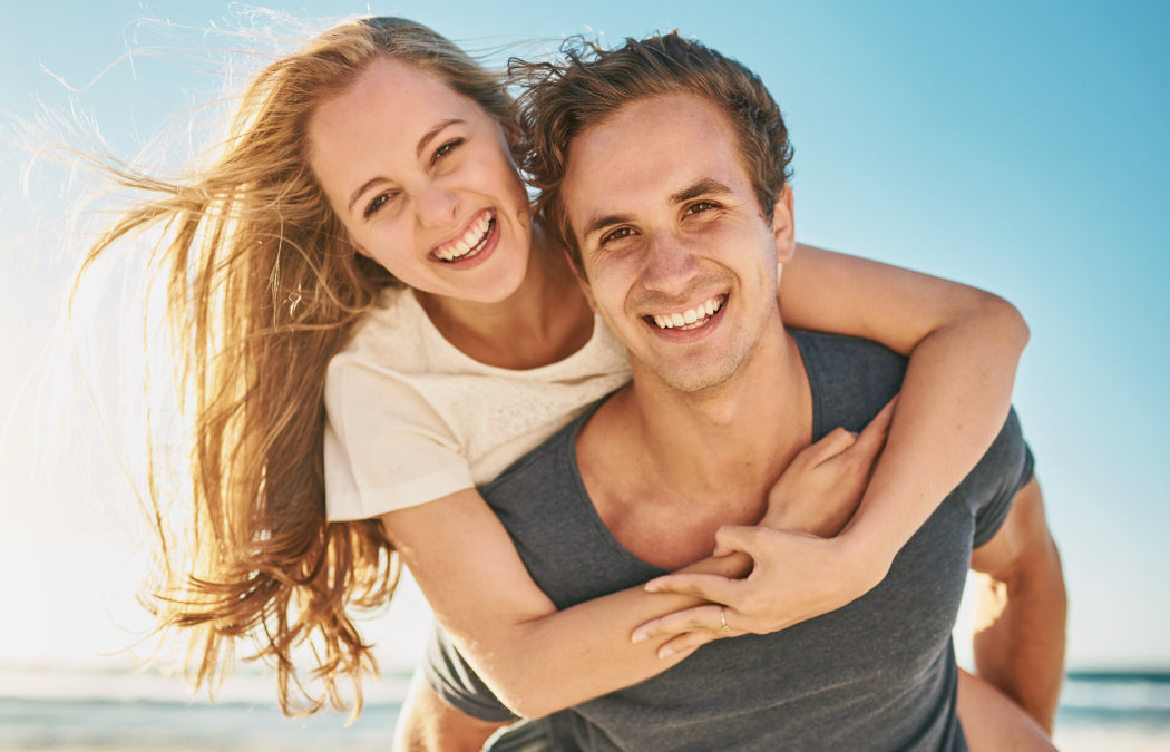 A woman piggybacks on a man at the beach; both are smiling and looking at the camera under a clear blue sky.