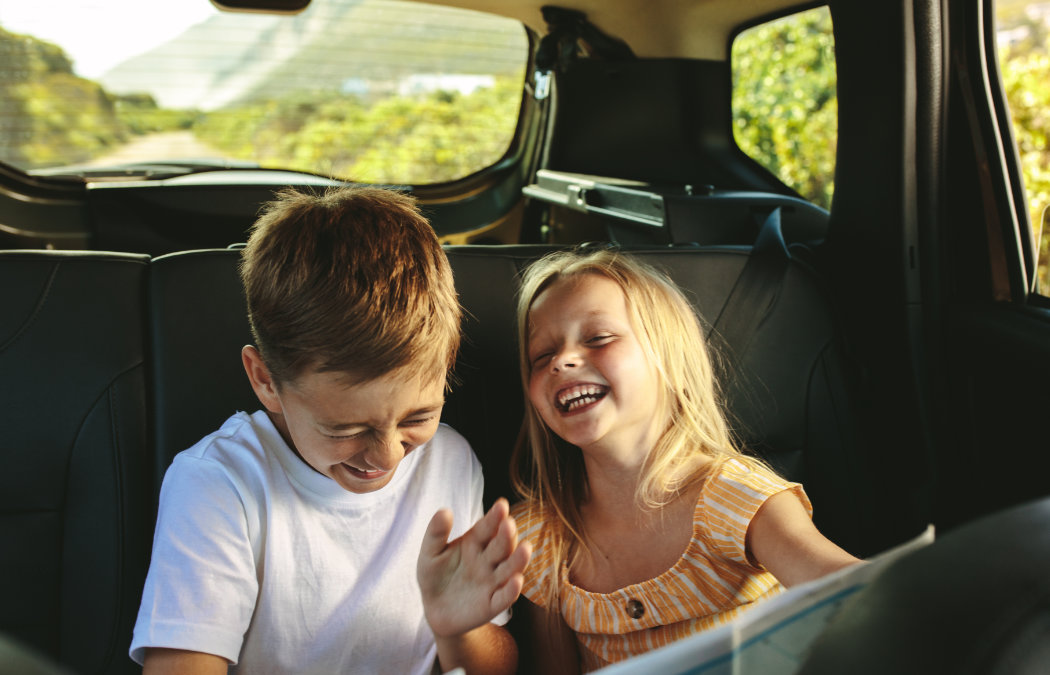 Two children sit in the backseat of a car, laughing and smiling, with greenery visible through the rear window.