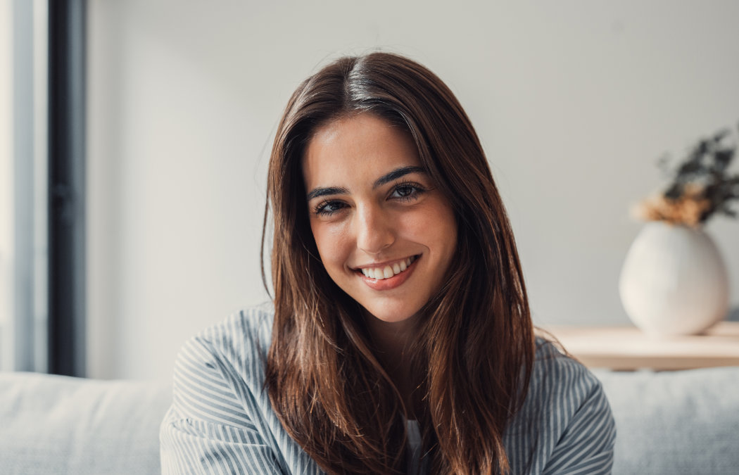 A young woman with long brown hair sits indoors, smiling at the camera. There is a white vase with dried flowers in the blurred background.